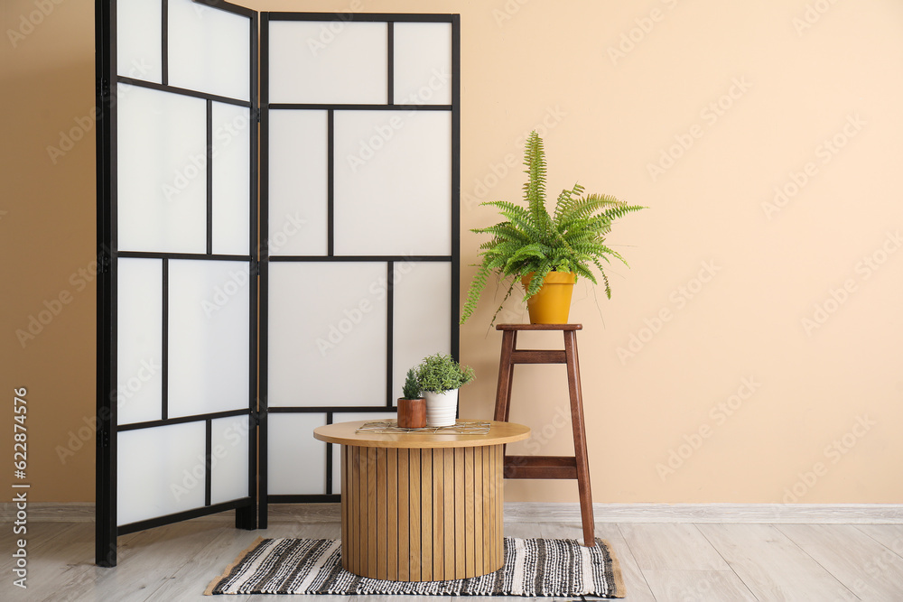 Coffee table, folding screen and houseplants near beige wall