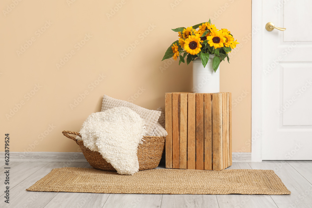 Vase with beautiful sunflowers and on wooden box and cushions in wicker basket near beige wall