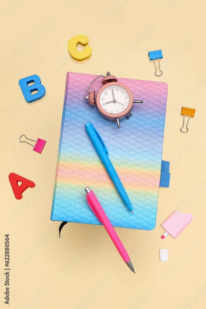 Notebook with different stationery and alarm clock on pale yellow background