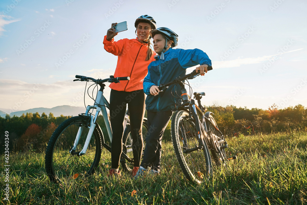 Mother and son ride a bike in park on green meadow in autumn day at sunset time. Family weekend. Wom