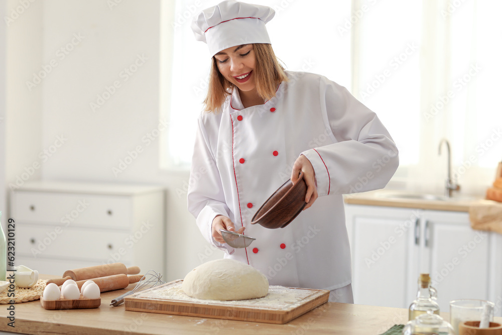 Female baker sprinkling dough with flour at table in kitchen