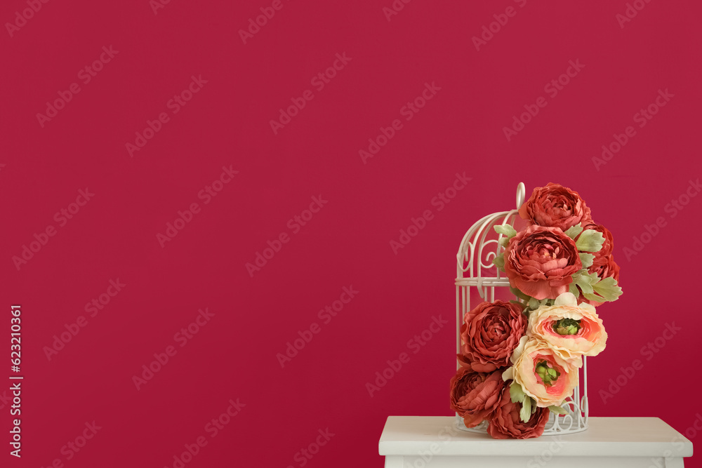 Cage with beautiful flowers on stool near pink wall