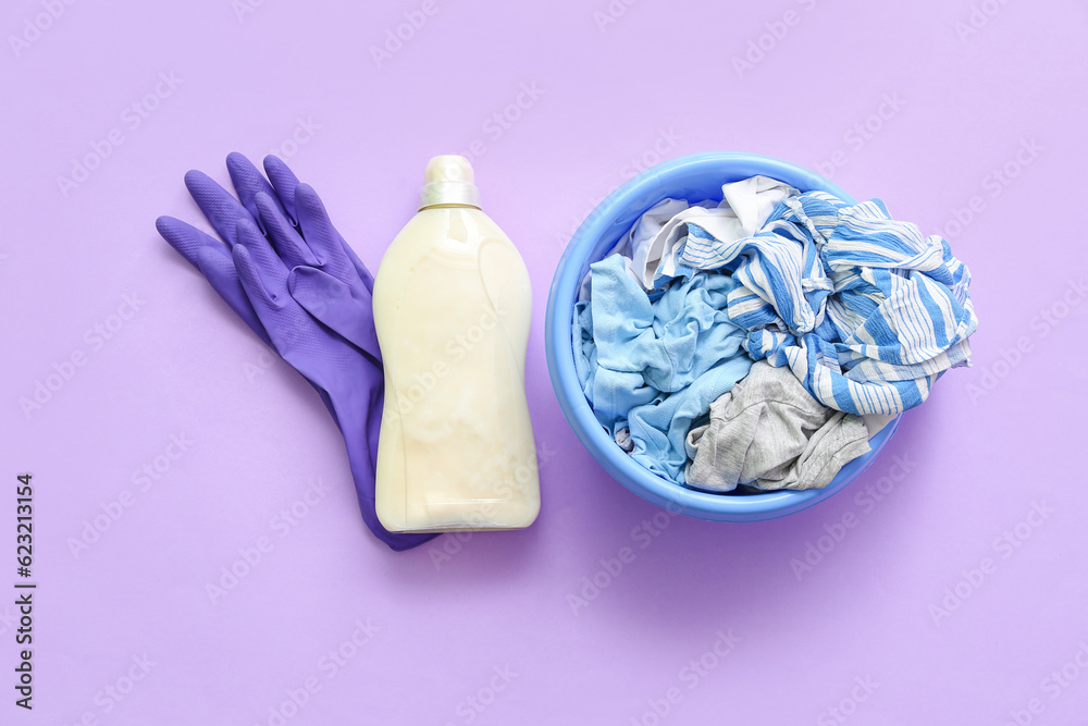 Plastic basin with dirty clothes, rubber gloves and bottle of laundry detergent on purple background