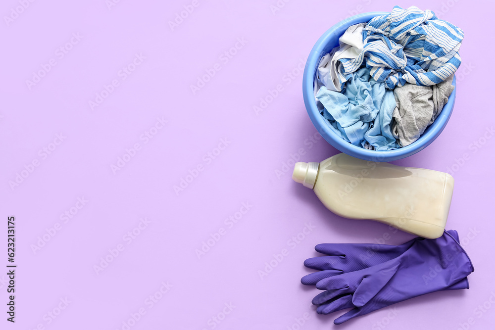 Plastic basin with dirty clothes, rubber gloves and bottle of laundry detergent on purple background