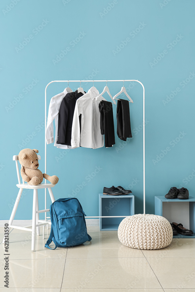 Stylish school uniform hanging on rack, backpack, shoes and pouf near blue wall in room