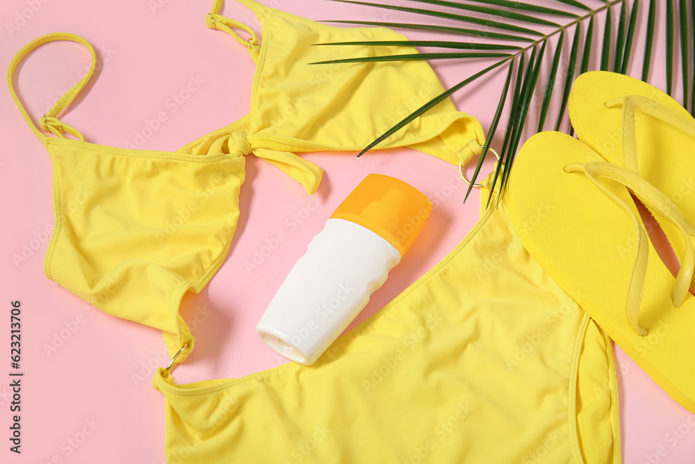 Stylish swimsuit, flip-flops, bottle of sunscreen cream and palm leaf on pink background, closeup