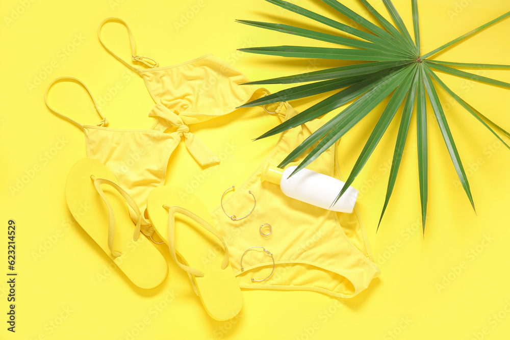 Stylish swimsuit, flip-flops, accessories, bottle of sunscreen cream and palm leaf on yellow backgro