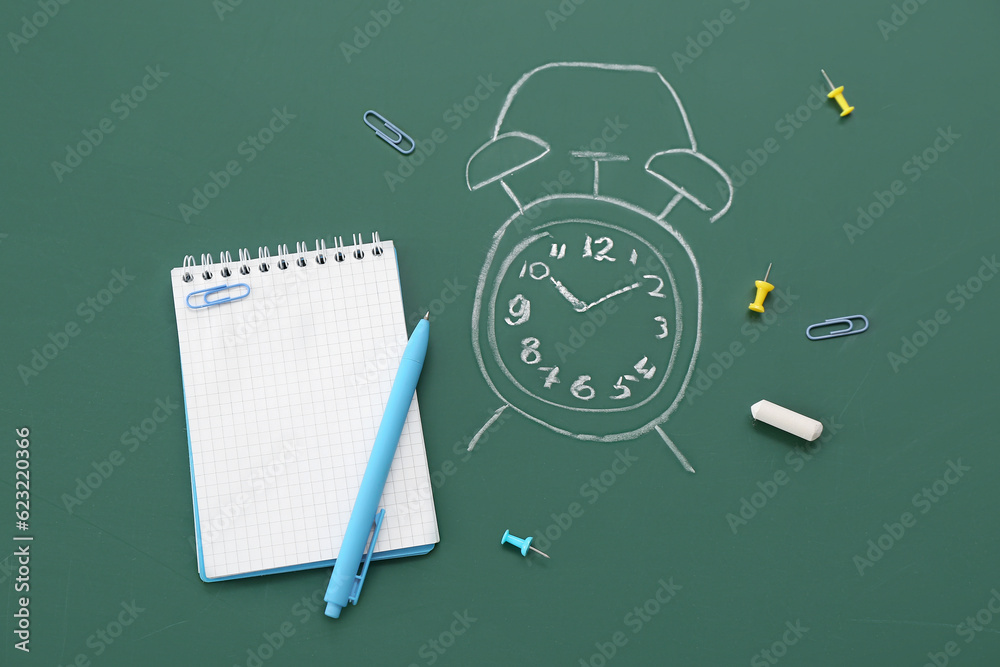 Blank notebook with stationery and drawn alarm clock on green chalkboard