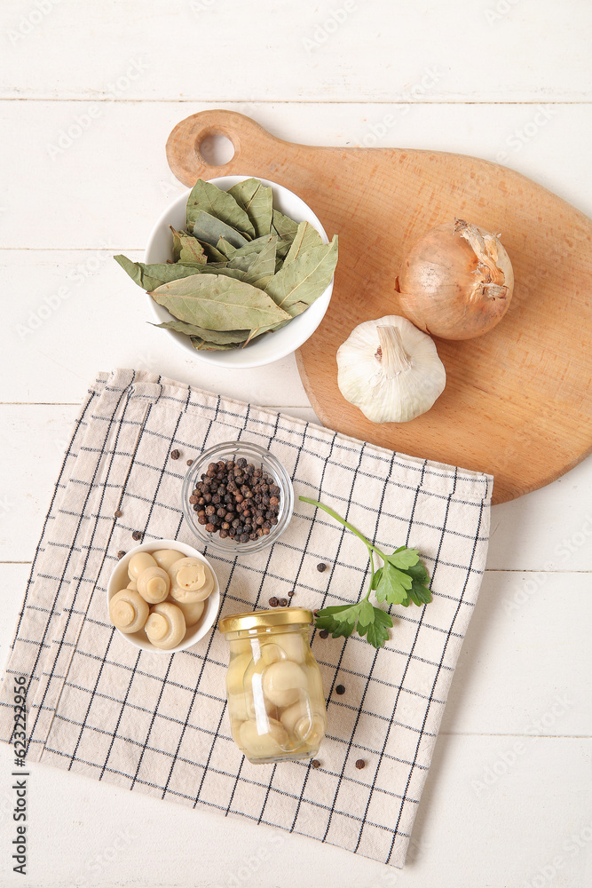 Jar and bowl of canned mushrooms with ingredients for preservation on white wooden background