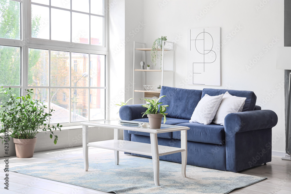 Interior of light living room with cozy blue sofa and modern laptop on table near big window