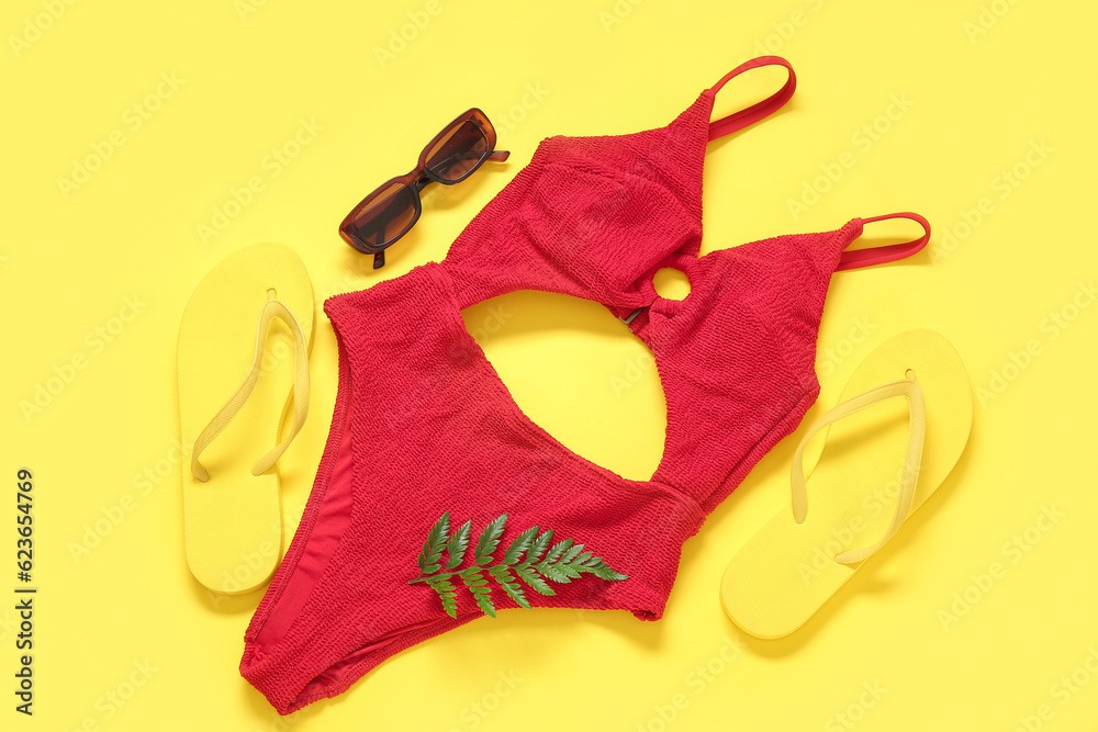 Stylish swimsuit, flip-flops, sunglasses and fern leaf on yellow background