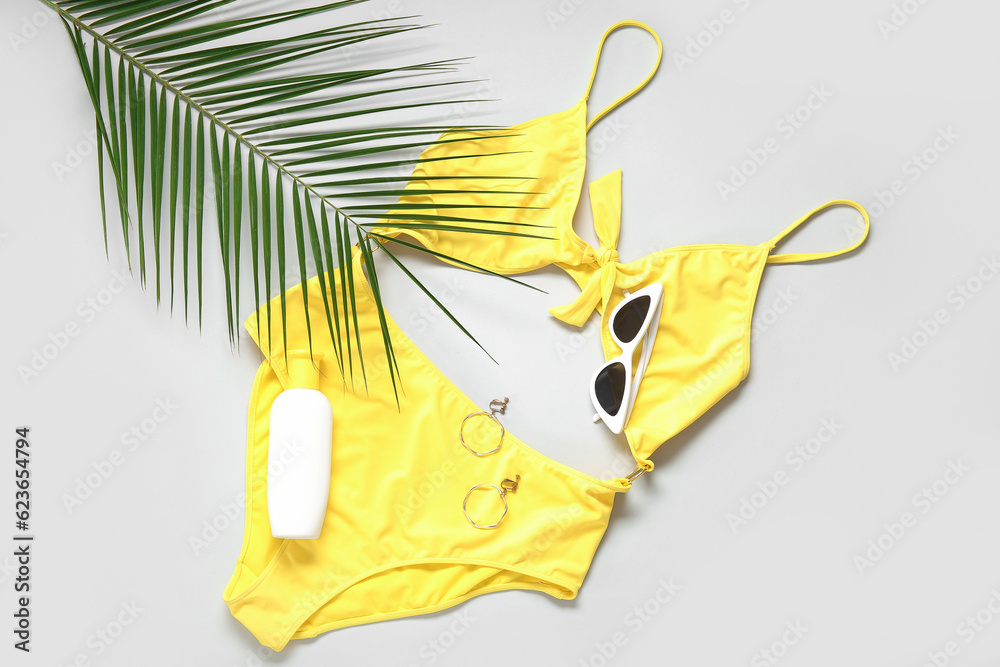 Stylish swimsuit, sunglasses, bottle of sunscreen cream, earrings and palm leaf on light background