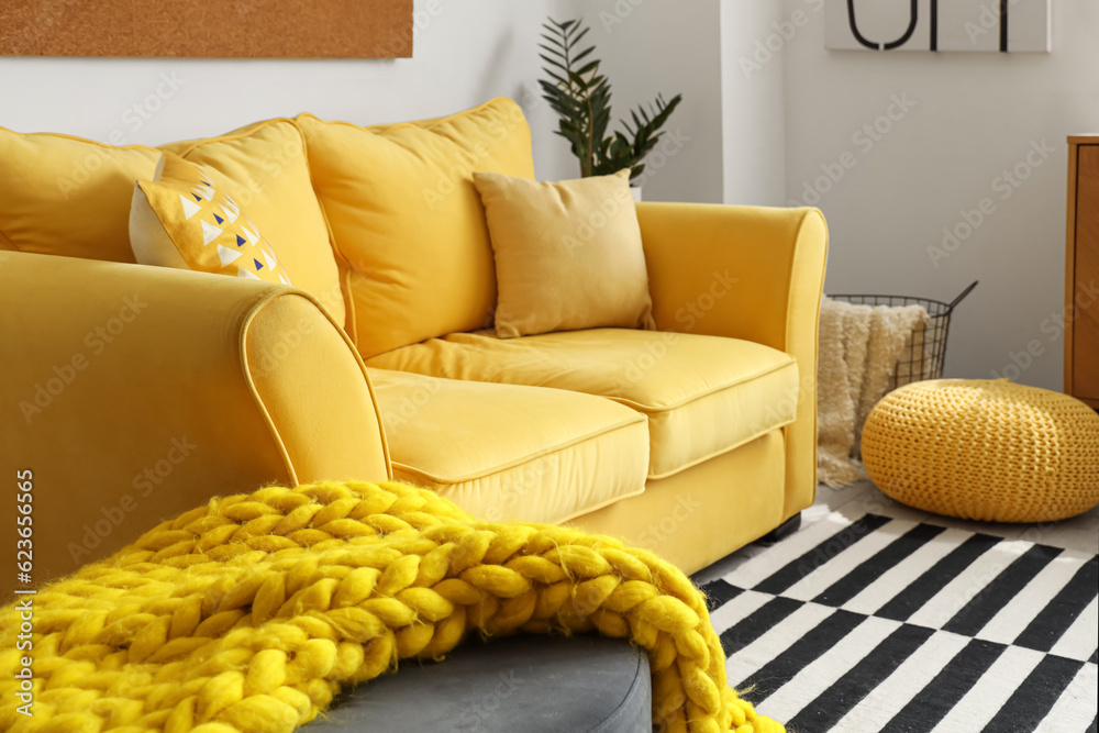 Interior of stylish living room with cozy yellow sofa and soft knitted blanket on pouf