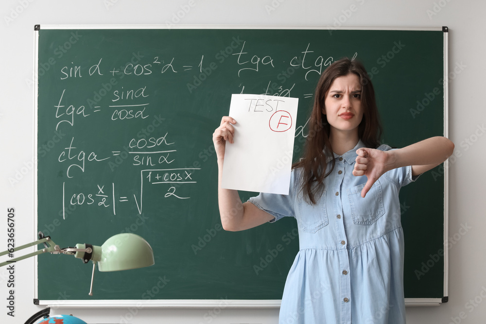 Female teacher with checked test sheet showing thumb-down near blackboard in classroom