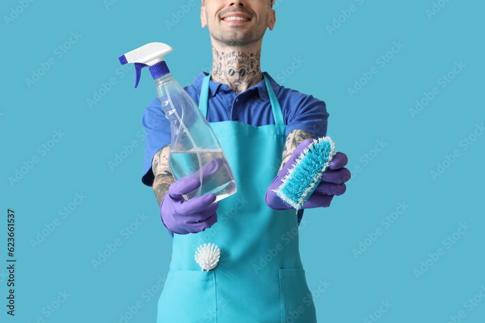 Young tattooed man with bottle of detergent and cleaning brush on blue background, closeup