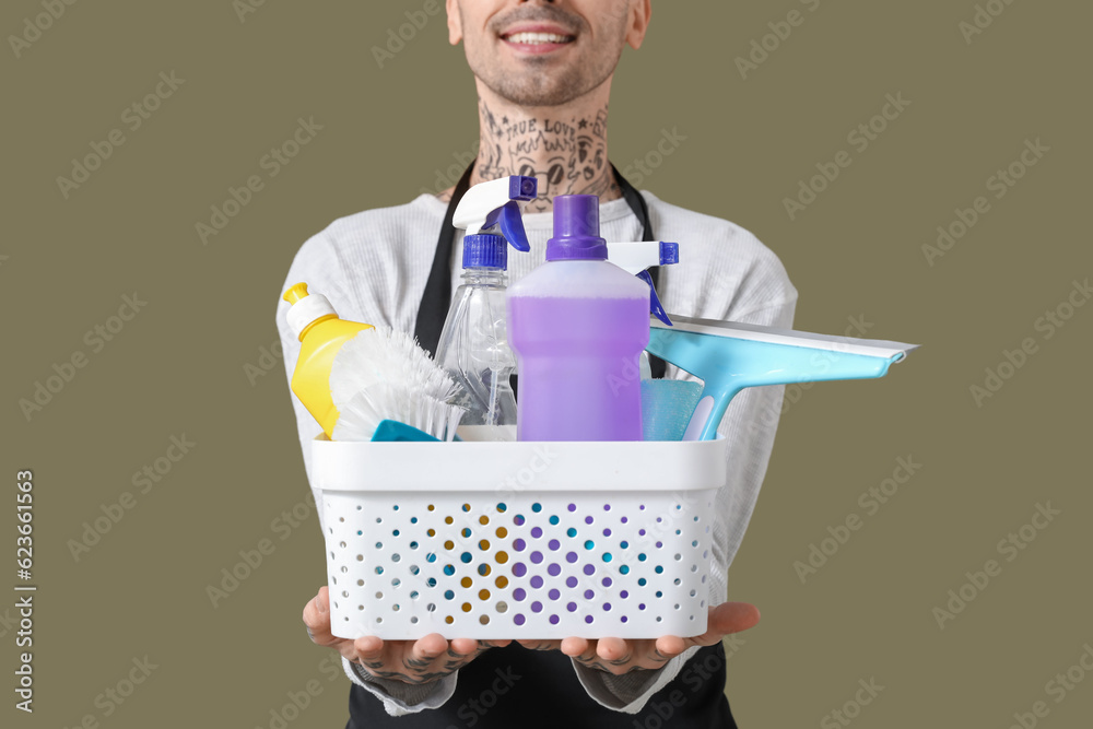 Young tattooed man with bucket of cleaning supplies on green background, closeup