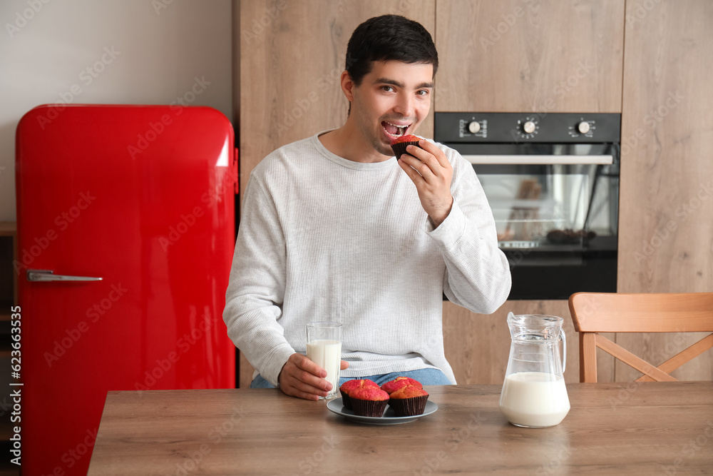 Young man with glass of milk eating muffin in kitchen