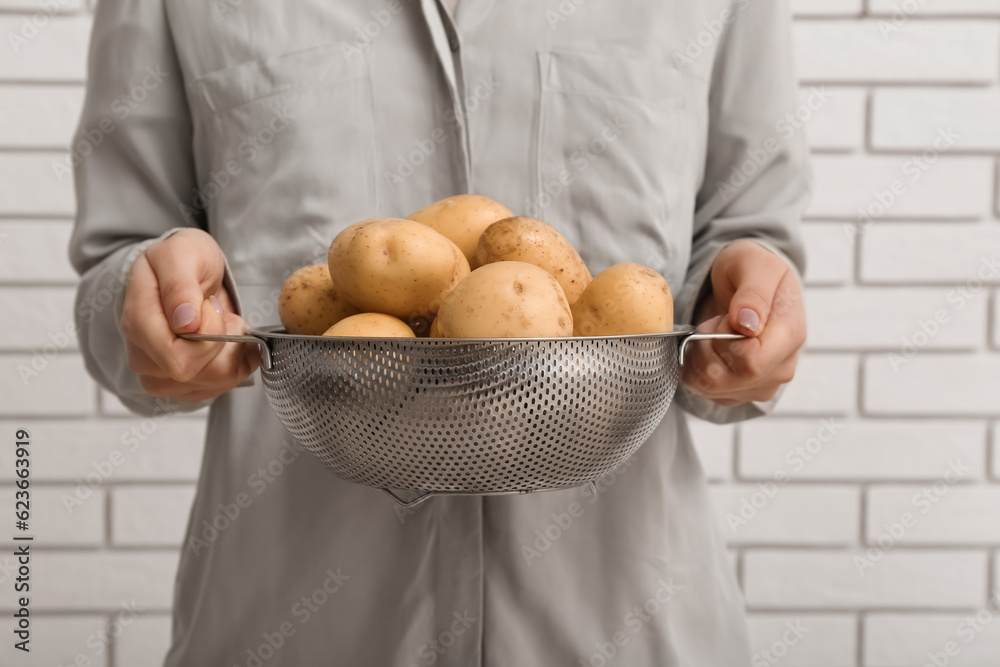 Woman holding colander with raw potatoes near white brick wall