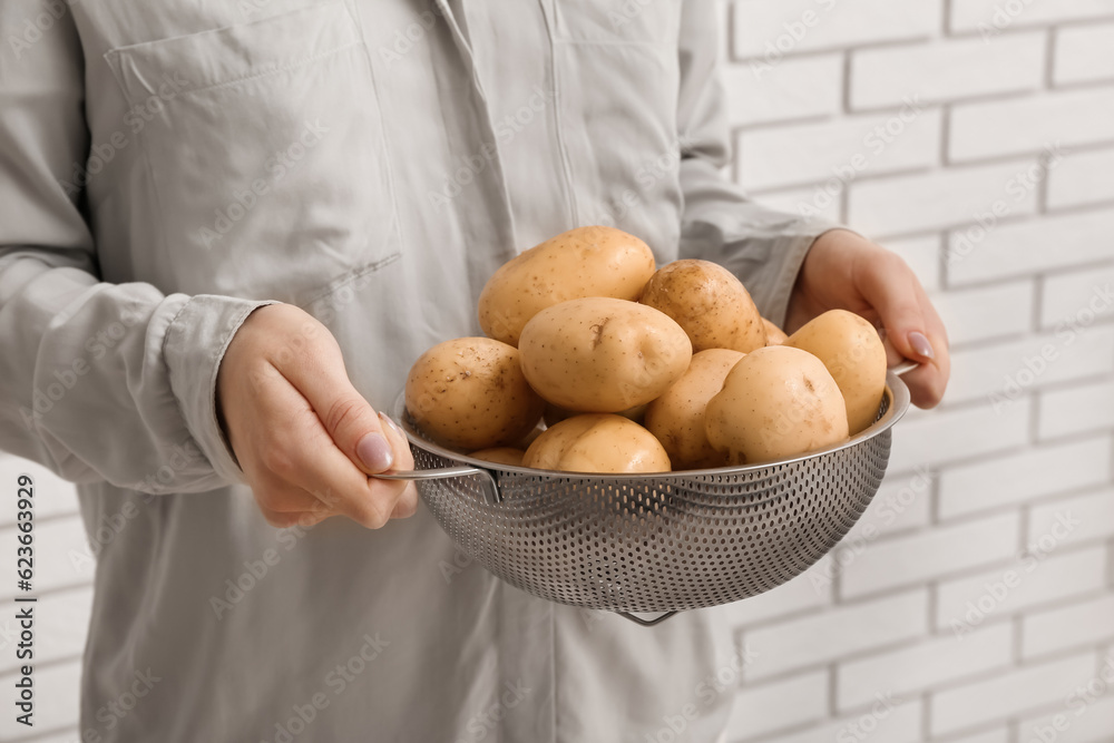 Woman holding colander with raw potatoes near white brick wall