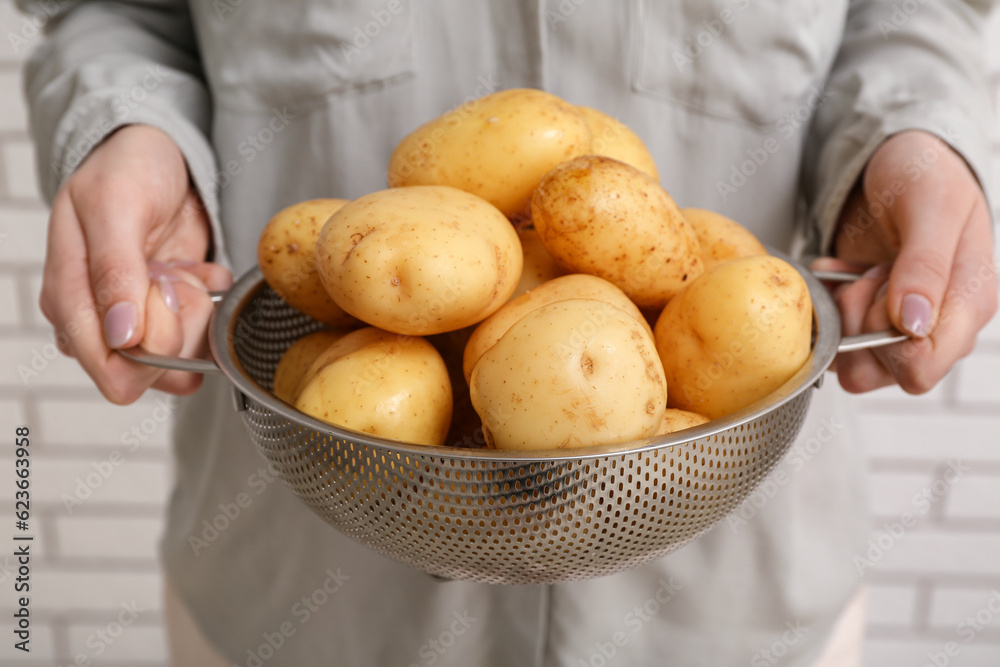 Woman holding colander with raw potatoes near white brick wall