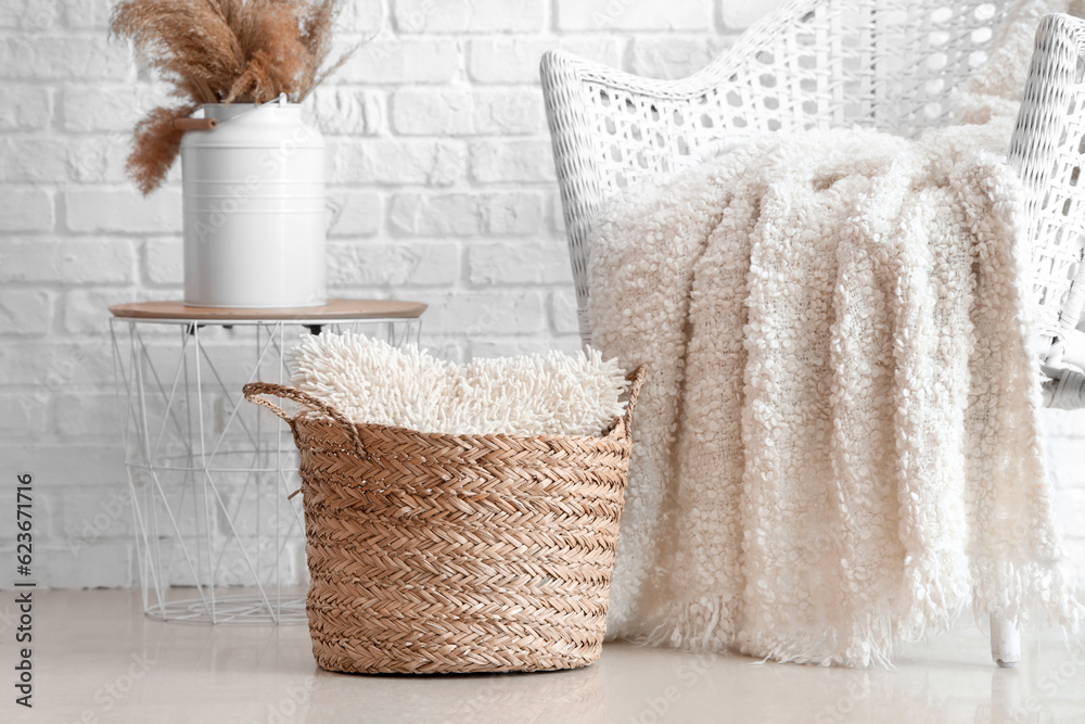 Rattan armchair with knitted plaid and wicker basket with cushion near white brick wall