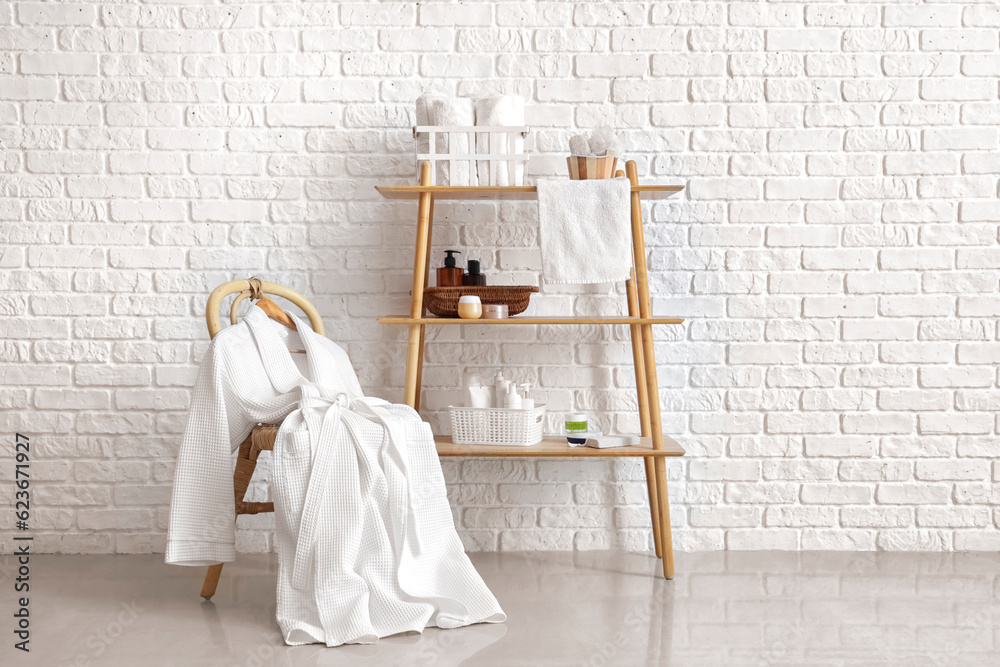 Shelving unit with bath accessories and bathrobe on chair near white brick wall