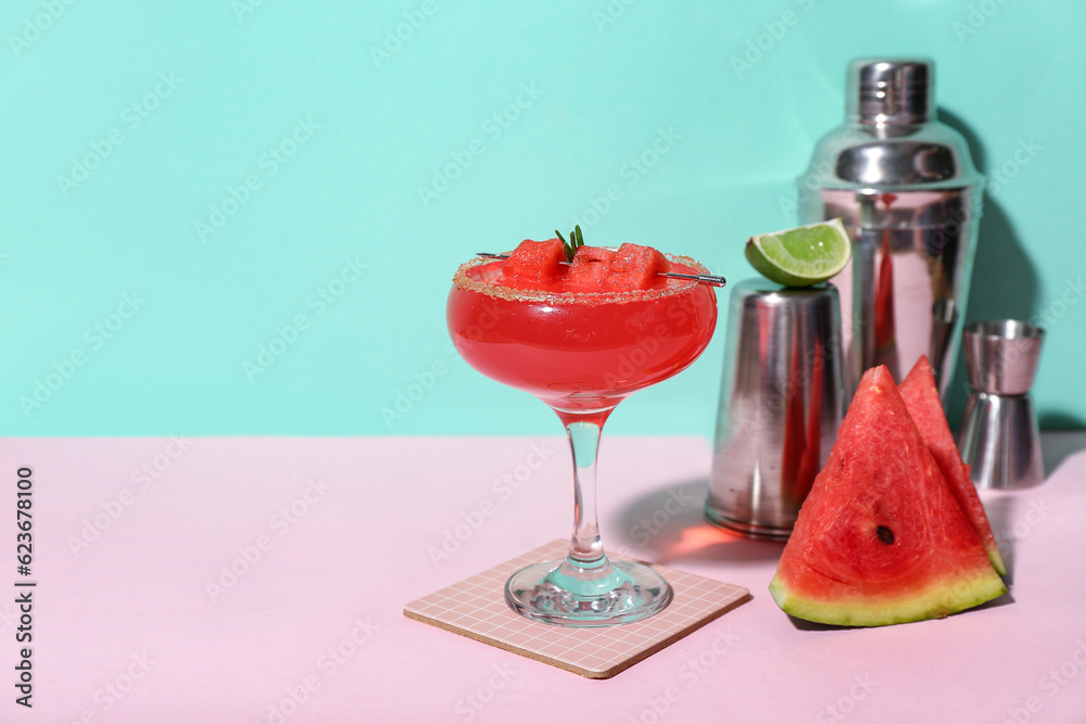 Glass of tasty watermelon cocktail, shakers and jigger on pink table near turquoise wall