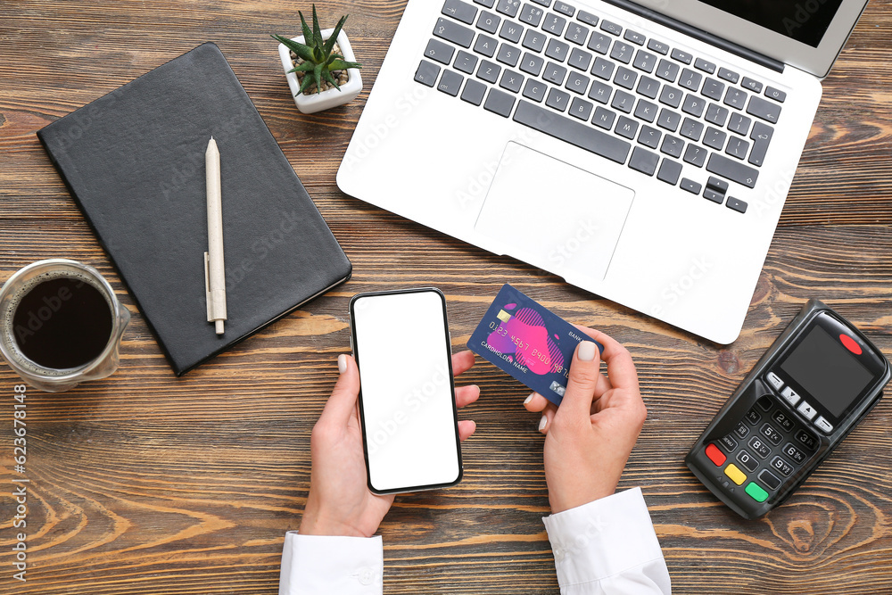 Female hand with payment terminal, mobile phone and credit card on wooden background