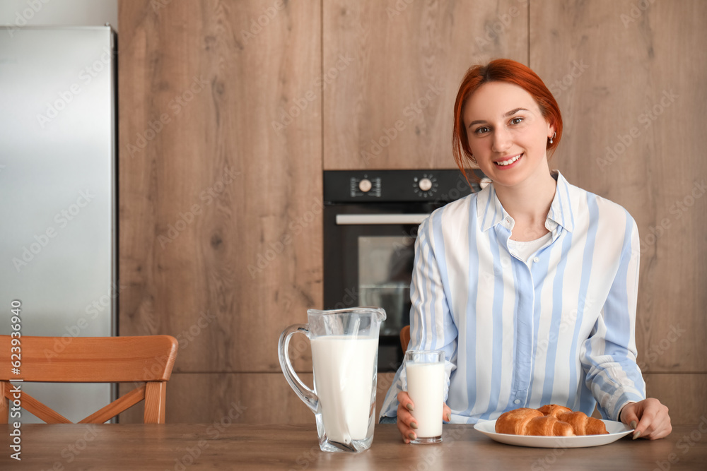 Young redhead woman with glass of milk and croissant in kitchen