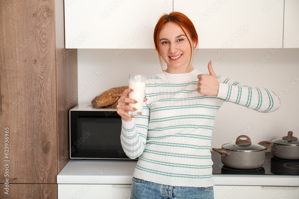 Young redhead woman with glass of milk showing thumb-up in kitchen