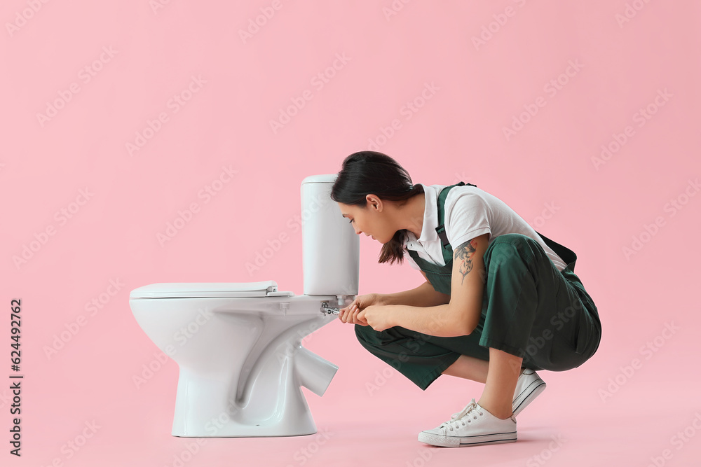 Female plumber repairing toilet bowl on pink background