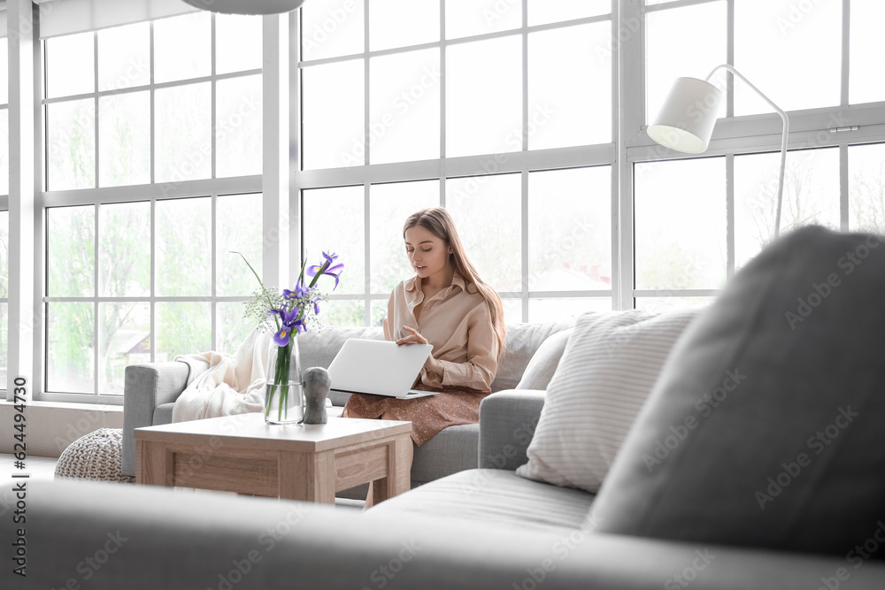 Pretty young woman sitting on grey sofa and using modern laptop in light living room