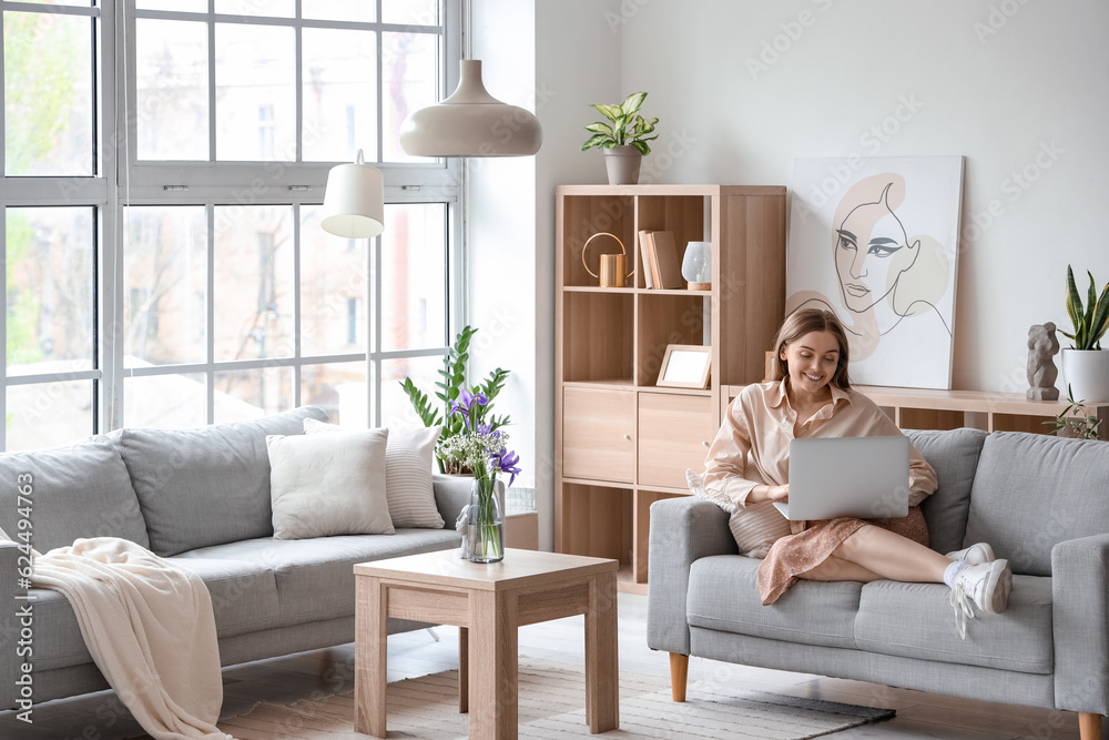 Pretty young woman sitting on grey sofa and using laptop in light living room