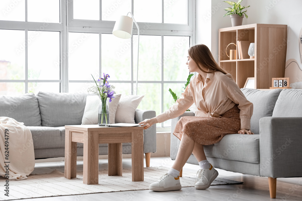 Pretty young woman sitting on grey sofa and taking modern laptop from table in light living room