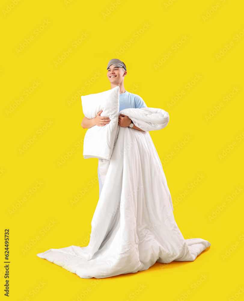 Young man with sleep mask, pillow and blanket on yellow background