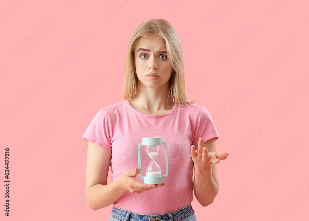 Stressed young woman with hourglass on pink background. Deadline concept