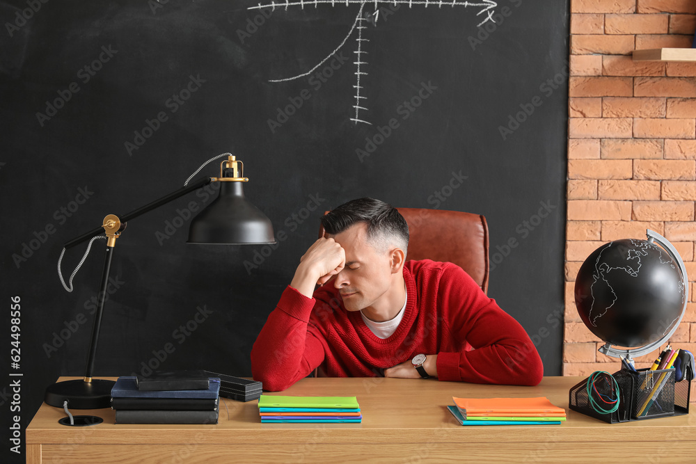 Stressed male teacher sitting at table in classroom