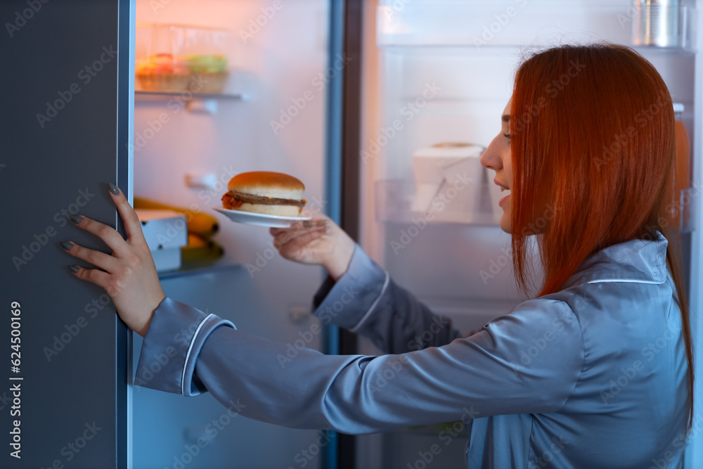 Young woman with tasty burger near open fridge in kitchen at night