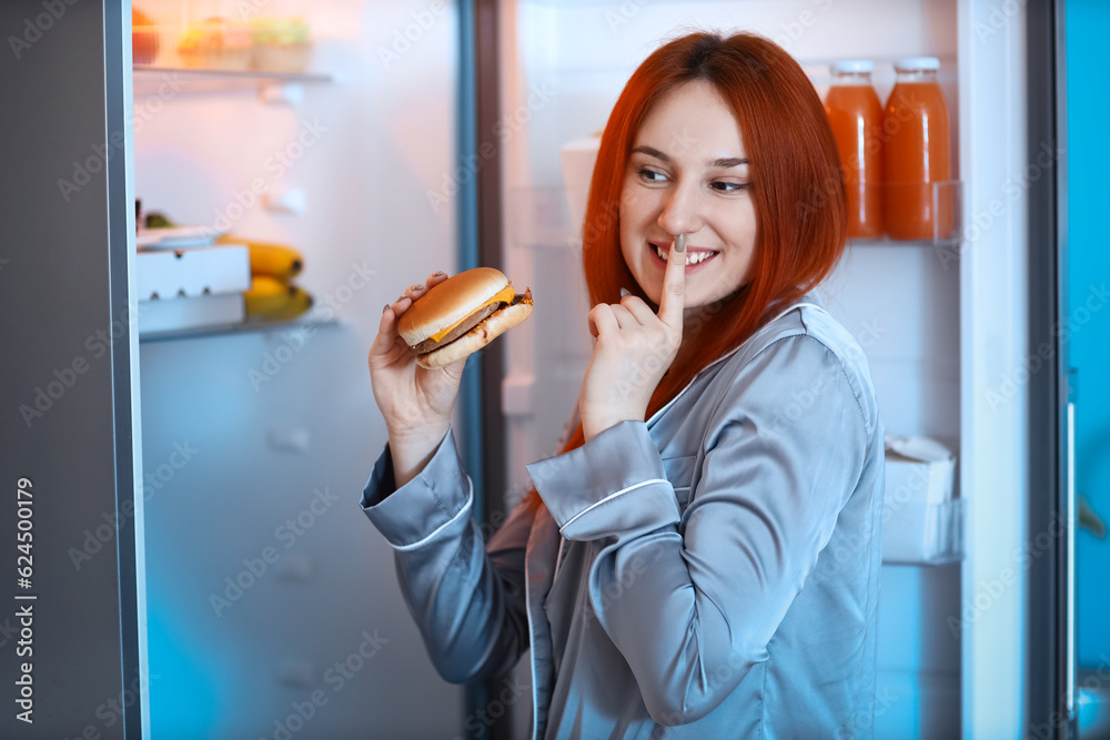 Young woman with tasty burger showing silence gesture near open fridge in kitchen at night