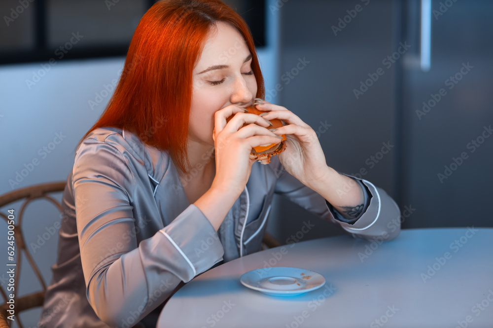 Young woman eating burger in kitchen at night