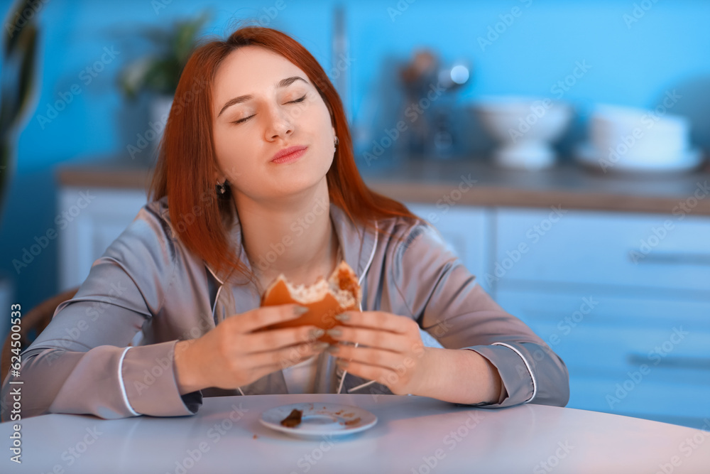 Young woman eating burger in kitchen at night