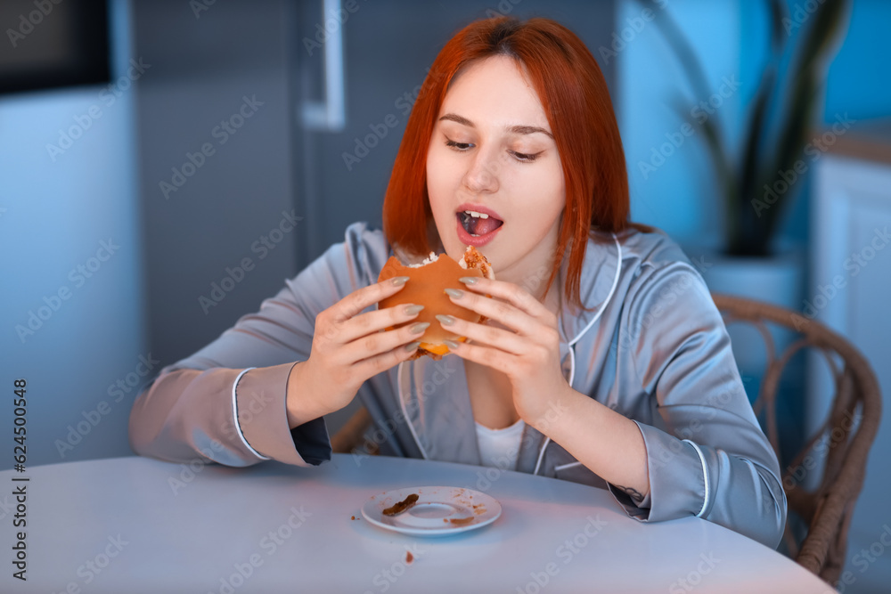 Young woman eating burger in kitchen at night