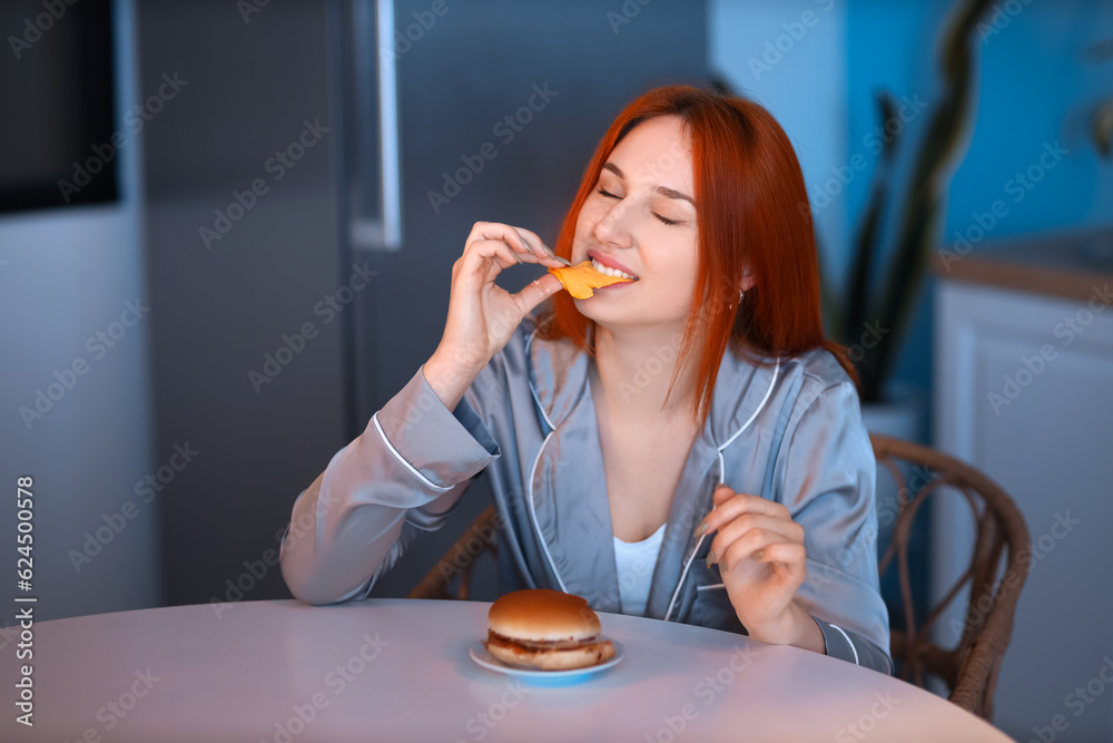 Young woman eating burger in kitchen at night