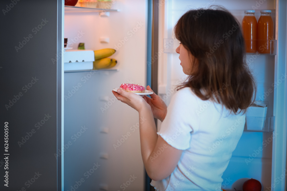 Young woman with tasty doughnut near open fridge in kitchen at night