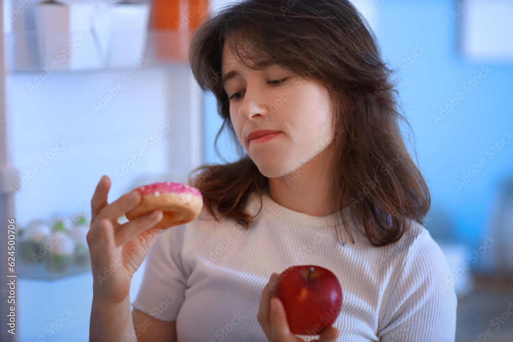 Young woman with doughnut and apple near open fridge in kitchen at night, closeup