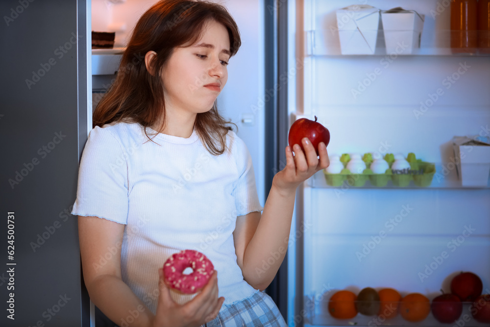 Young woman with doughnut and apple near open fridge in kitchen at night