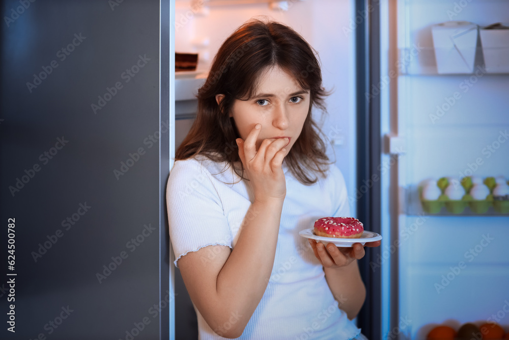 Young woman with tasty doughnut near open fridge in kitchen at night