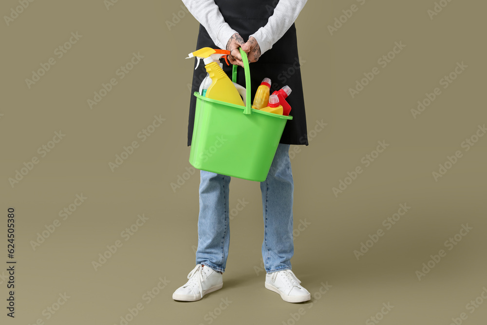 Young tattooed man with bucket of cleaning supplies on green background