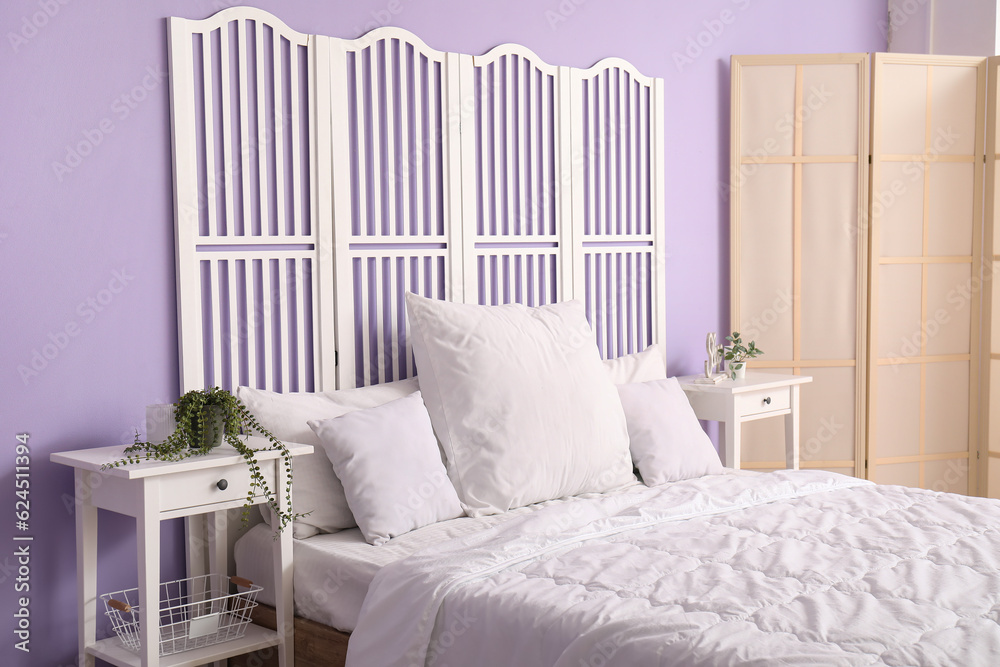 Interior of stylish bedroom with comfortable bed, white pillows, bedside tables and folding screen