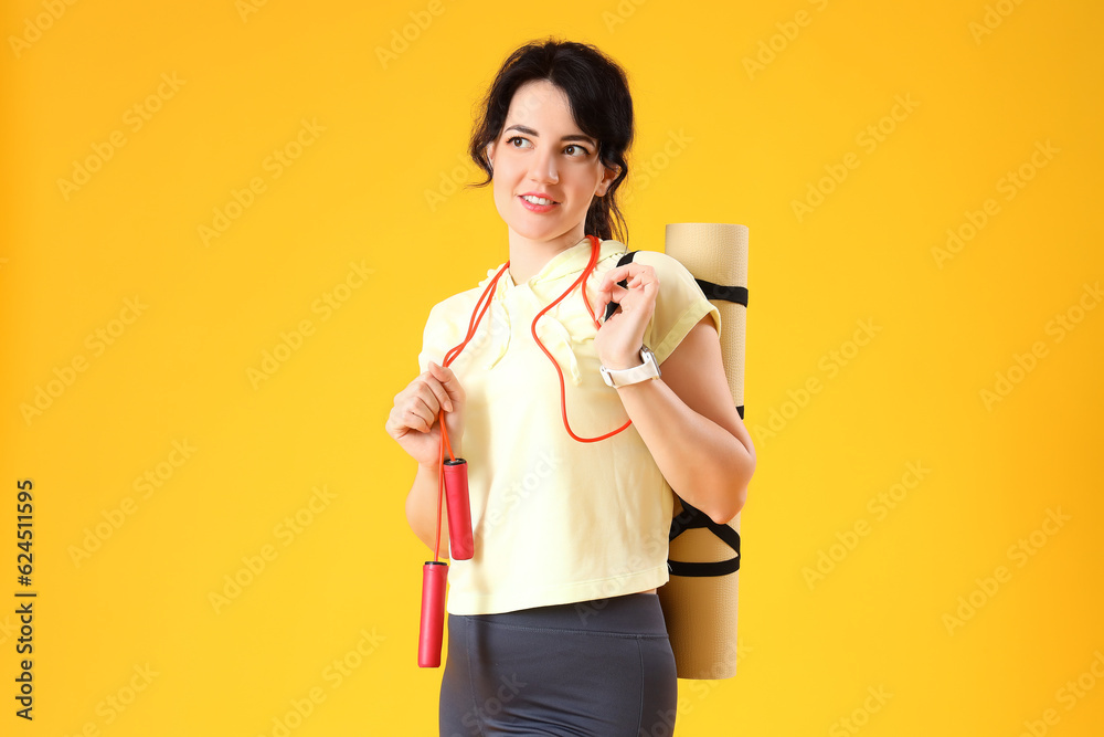 Sporty young woman with jumping rope and yoga mat on yellow background